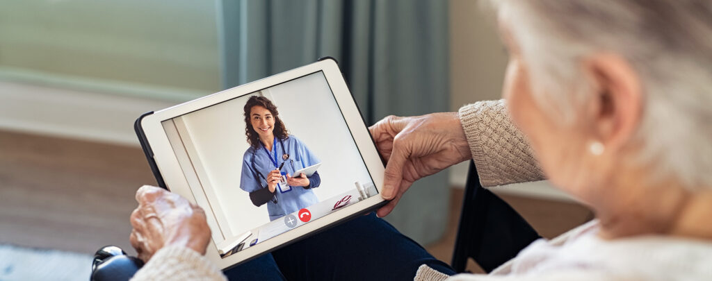 Back view of senior woman sitting in wheelchair making video call with her doctor while staying at home during covid pandemic. Close up of helpful general practitioner in video conference with old woman on digital tablet. Sick woman in online consultation from home: distance and telehealth concept.